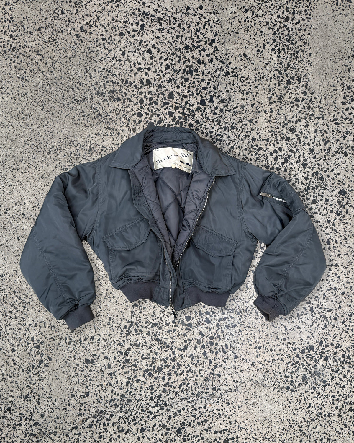 Charcoal Capsule Bomber, laid flat, showing the collar, front pockets and ribbed hem.