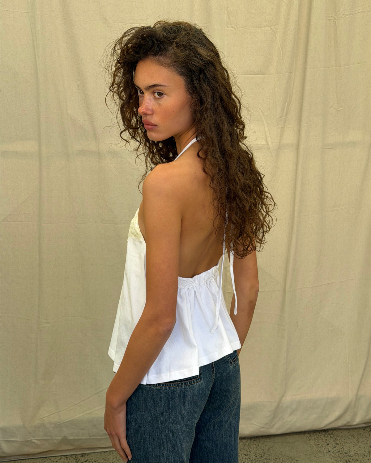 model:XS/6AU
Back view of the white Jersey Lace Halter Top, showing its open back, halter ties and relaxed hem over blue jeans.