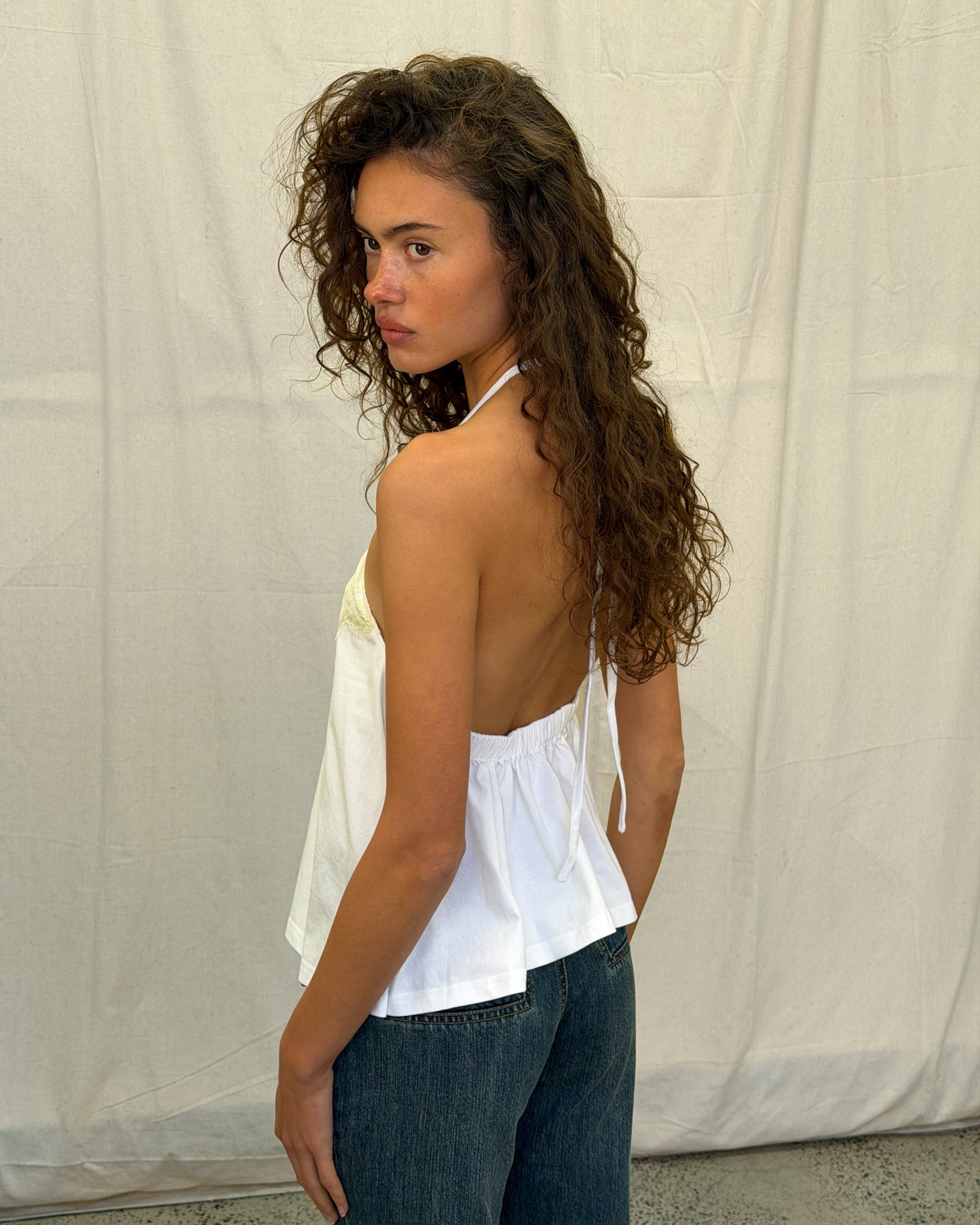 model:XS/6AU H:179CM
Side view of the white Jersey Lace Halter Top, showing the lace neckline, open back and loose hem with grey shorts.
