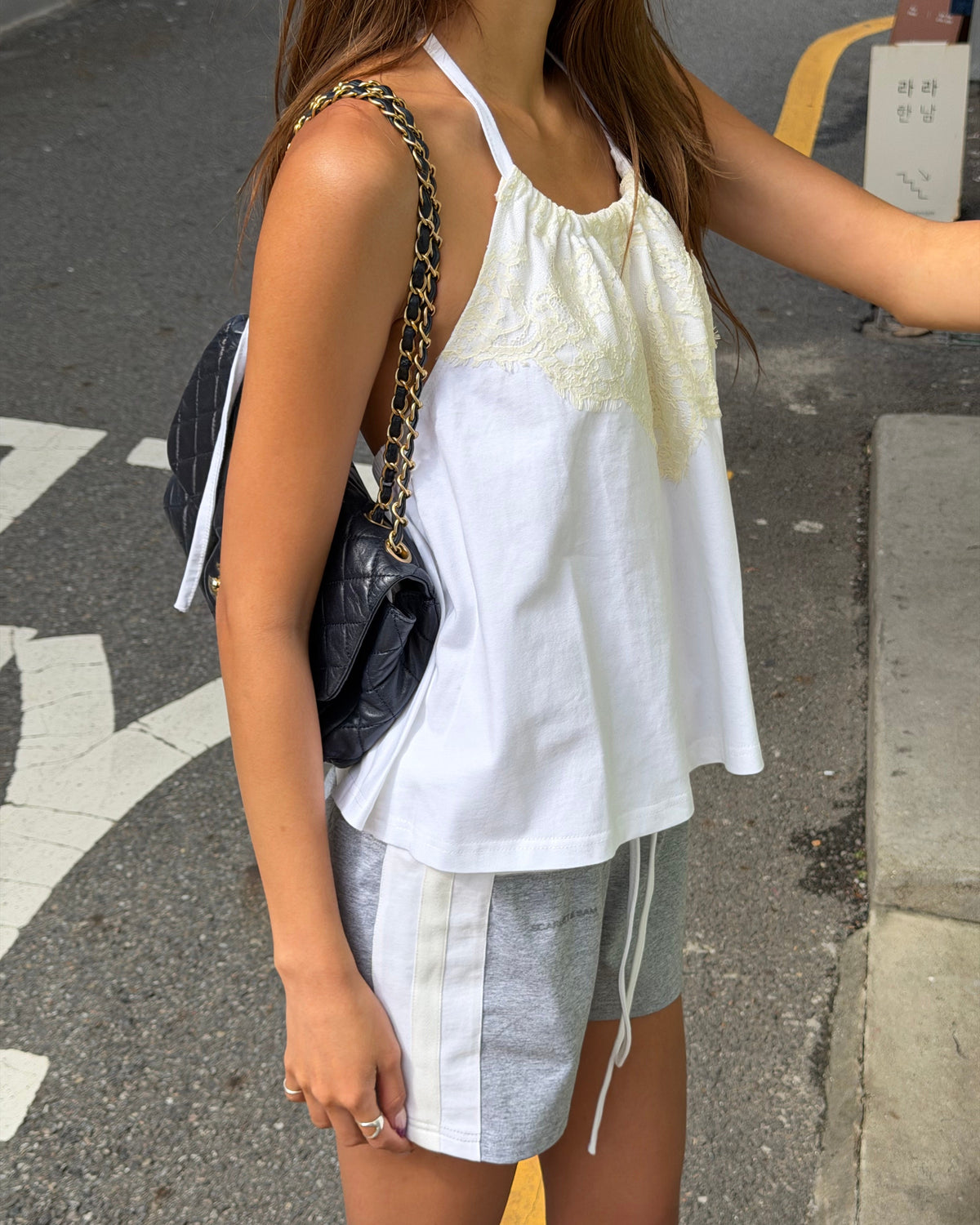 model:XS/6AU
Side view of the white Jersey Lace Halter Top, showing the lace neckline, open back and loose hem with grey shorts.