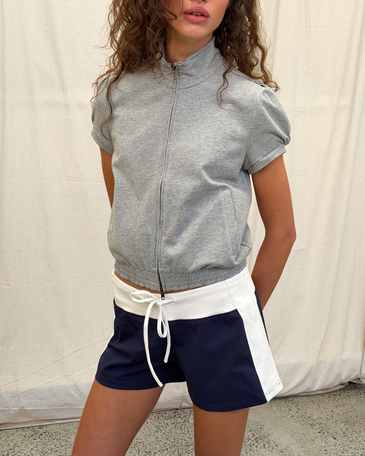 model:S/8AU H:179CM 
Grey Jersey Zip Top, front close-up on a model, showing the zipped high collar, puff sleeves and gathered hem with navy Jersey Mini Shorts.