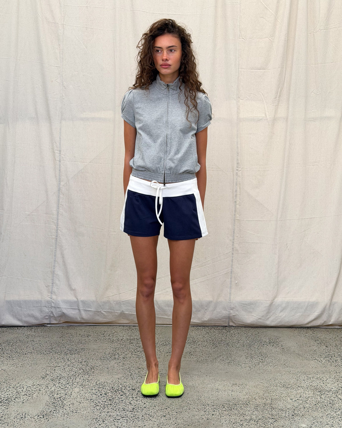 model:S/8AU H:179CM 
Navy Jersey Mini Short in a full-length front view, styled with a grey Jersey Zip Top and highlighter-yellow ballet flats.