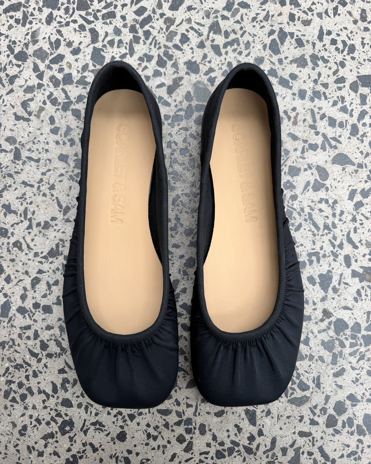 Black Noa Neoprene Ballet Flats laid on terrazzo, showing the gathered square toes and tan insoles from above.