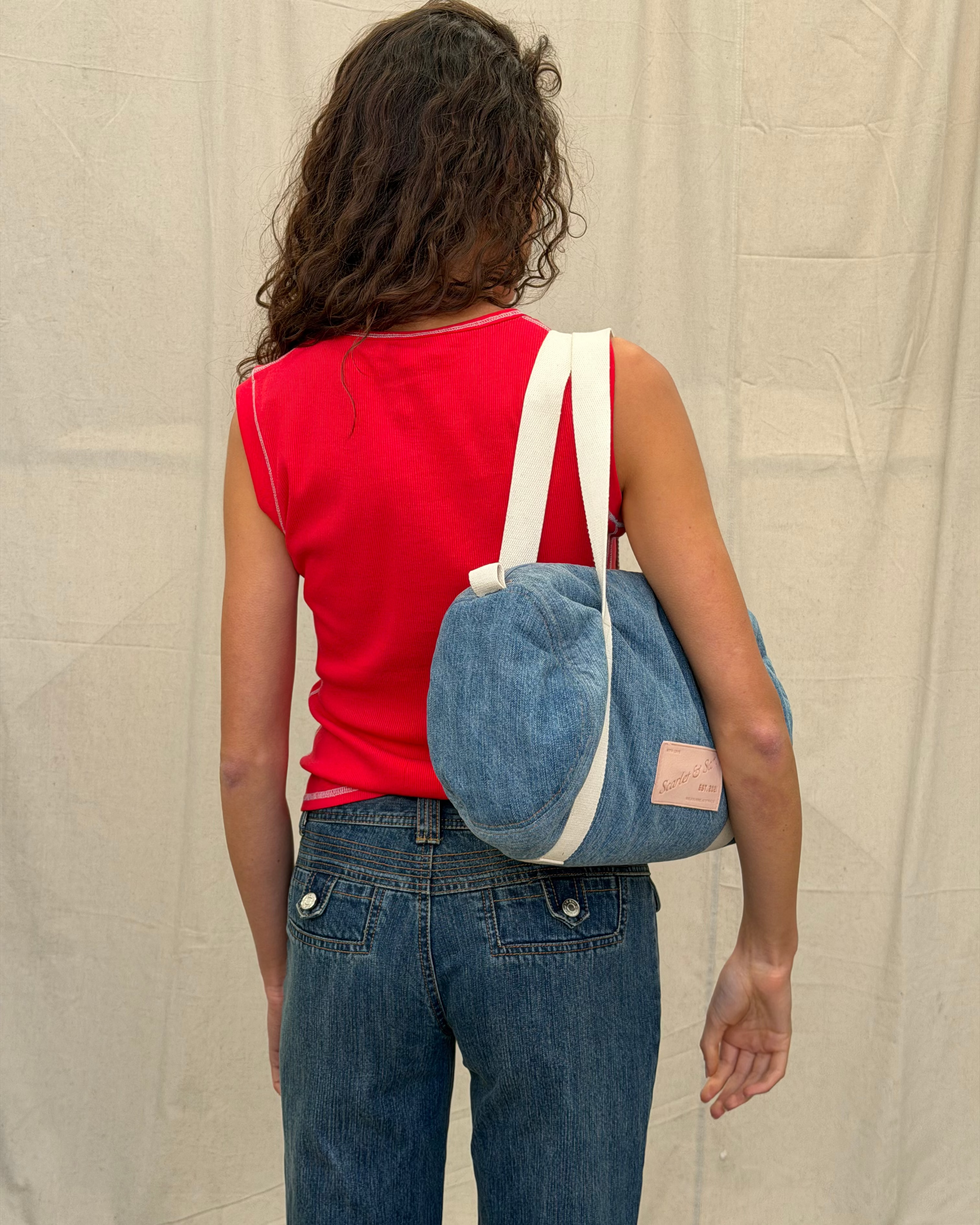 Denim Padded Barrel Tote worn over the shoulder, showing the rounded side, cream straps and front logo patch.