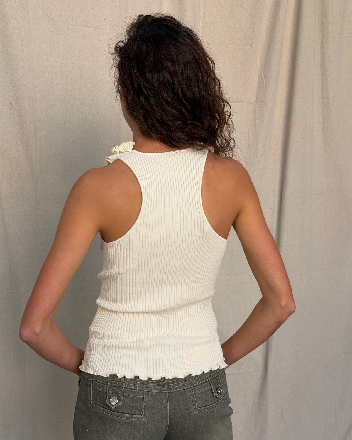 model:XS/6AU H:179CM
Back view of the cream Ruffle Knit Tank, showing the racerback cut, ribbed texture and ruffled hem with khaki jeans.