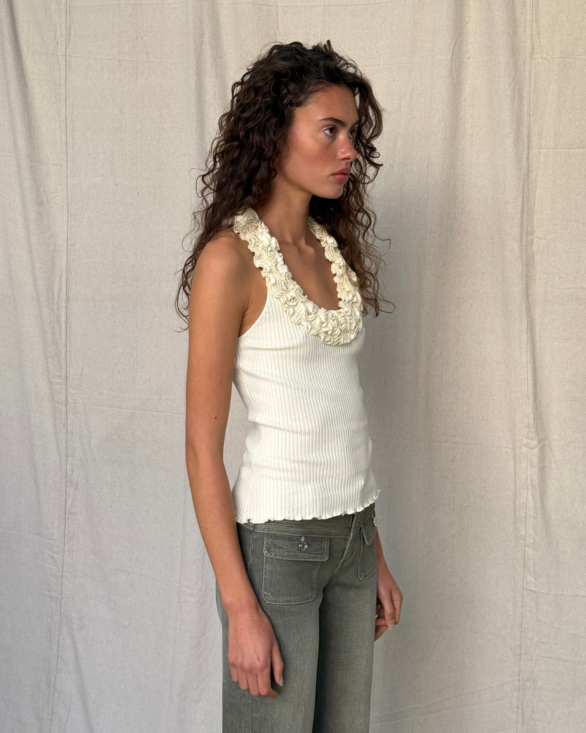 model:XS/6AU H:179CM
Three-quarter view of the cream Ruffle Knit Tank, showing its fitted ribbed shape and ruffled neckline with khaki jeans.